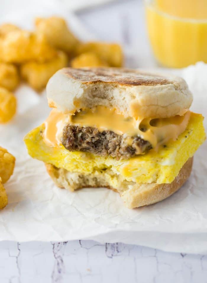 Crisp and buttery English muffins topped with fluffy eggs, bacon, sausage, and cheese. Easy to freeze ahead of time and heat in just 3 minutes! A sausage egg and cheese Breakfast Sandwiches with a bite taken out of it and tater tots in the background.