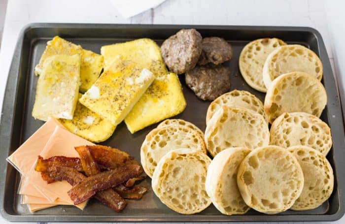 Crisp and buttery English muffins topped with fluffy eggs, bacon, sausage, and cheese. Easy to freeze ahead of time and heat in just 3 minutes! breakfast ingredients on a baking tray for making freezer breakfast sandwiches