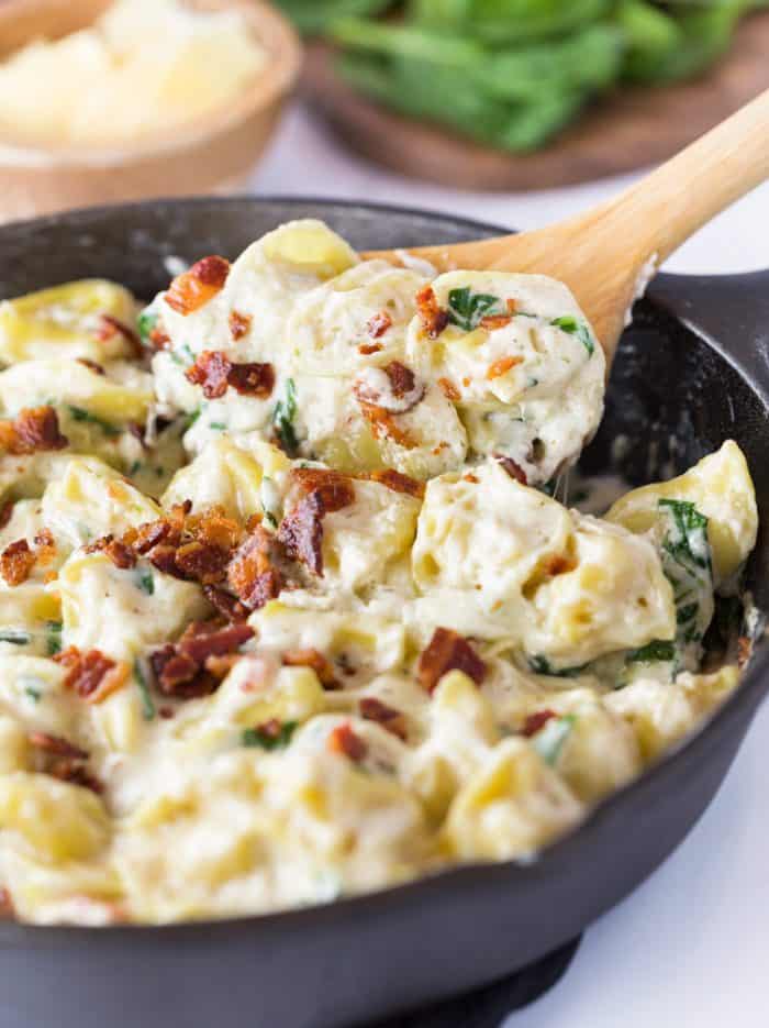 This Tortellini Al Forno features stuffed tortellini tossed in a rich and creamy garlic cheese sauce and topped with crumbled bacon. | The Cozy Cook | #Tortellini #Pasta #OliveGarden #AlForno #Dinner, #Cheese #Parmesan A wooden spoon scooping Tortellini Al Forno out from a cast iron skillet.
