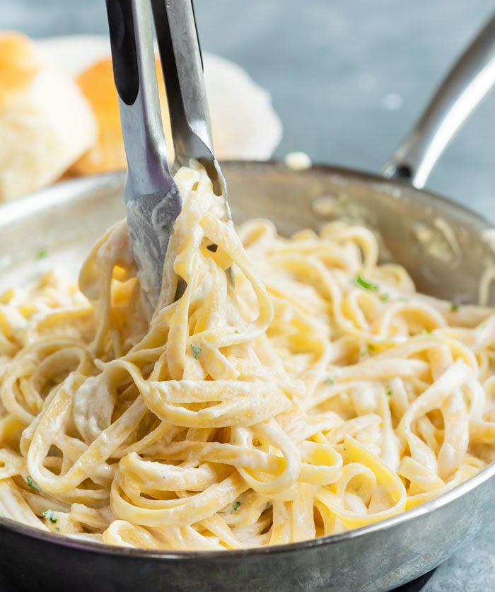 A skillet filled with Fettuccine Alfredo with kitchen tongs pulling noodles up.