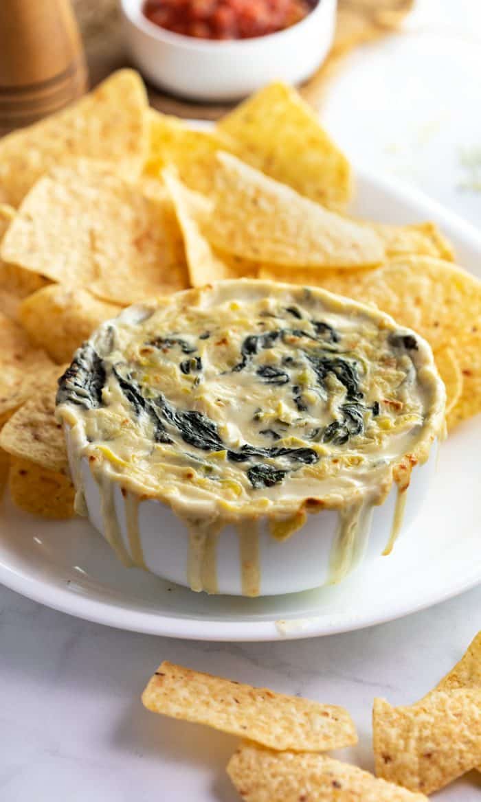 This ultra-creamy Spinach and Artichoke Dip tastes just like the Applebee's version! It's easy to make at home in under 30 minutes, just combine the ingredients until melted! This is perfect with tortilla chips, pita chips, crusty Artisan breads, and vegetables! | The Cozy Cook | #appetizer #dip #applebees #copycat #spinach #artichoke White bowl overflowing with spinach artichoke dip and corn tortillas on white plate