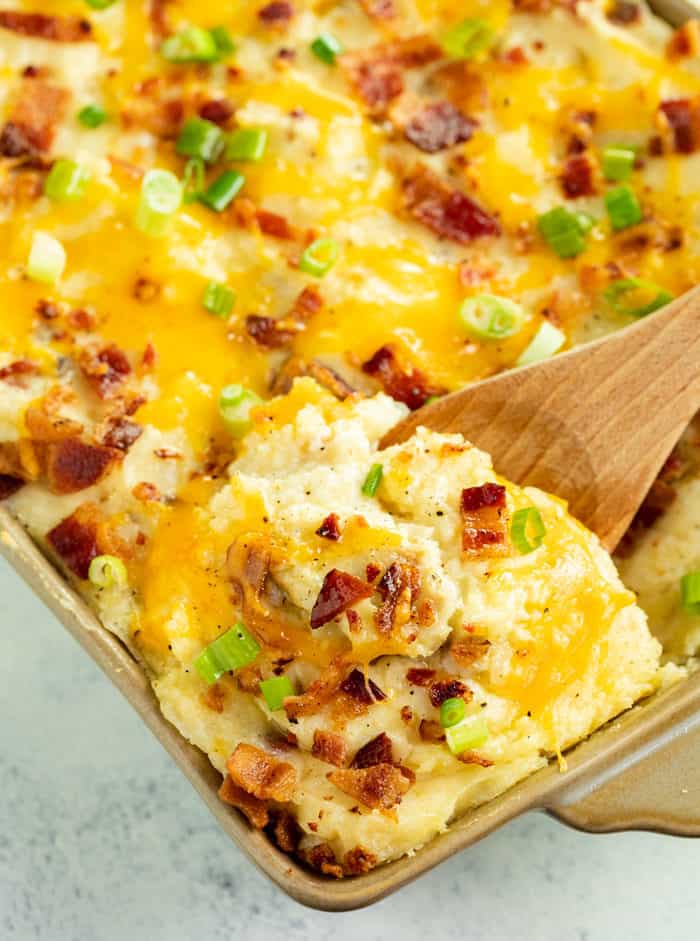This Twice Baked Potato Casserole recipe from The Pioneer Woman is an easy make ahead side dish idea for Thanksgiving, Christmas, or any family dinner! #comfortfood #bacon #potatorecipes #mashedpotatoes #sides #holidayrecipes A wooden spoon scooping out twice baked potato casserole topped with melted cheese, bacon, and green onions.