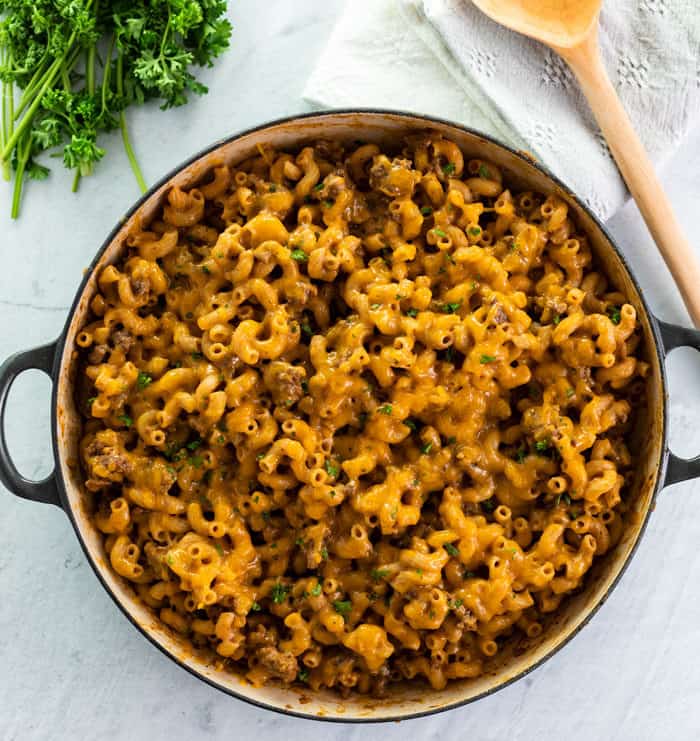 This Cheeseburger Casserole is made in one pot and takes just 30 minutes! It's a cheesy, meaty, and easy macaroni dinner. A homemade version of Hamburger Helper! #comfortfood #pasta #familydinneridea #dinnersforkids overhead view of a large skillet filled with warm cheeseburger casserole with a white background next to parsley and a wooden spoon.