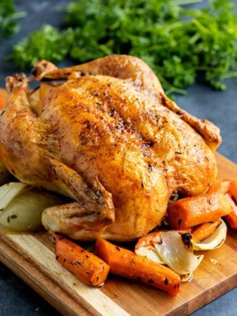 A roast chicken on a board with carrots and onions.