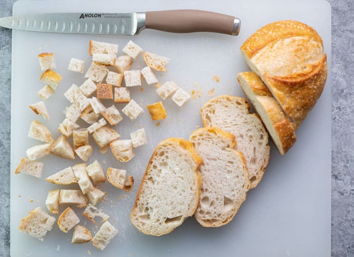 Learn how to make EASY homemade breadcrumbs! Store up to 3 months in the freezer and use for homemade meatballs, meatloaf, chicken, stuffing, casserole toppings, and more! #homemadebreadcrumbrecipe #easyrecipe #bread #breading #frying #panko A loaf of Italian bread being cut into cubes on a white cutting board.