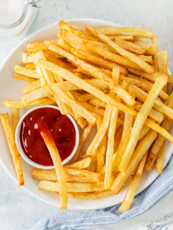 A plate of golden french fries on a white plate with a bowl of ketchup for dipping.