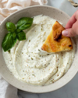 A bowl full of Feta Dip with a hand dipping bread into it with a garnish of basil.