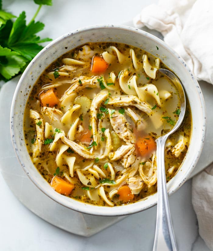 A bowl of chicken noodle soup with a spoon in the bowl and parsley.