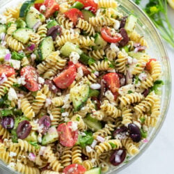 Greek Pasta Salad in a glass bowl with dressing, veggies, and Feta cheese.