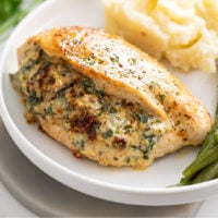Stuffed Chicken Breast on a white plate with green beans on the side and mashed potatoes in the back.