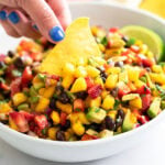 A hand dipping a tortilla chip into a white bowl of Mango Salsa.