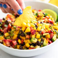 A hand dipping a tortilla chip into a white bowl of Mango Salsa.