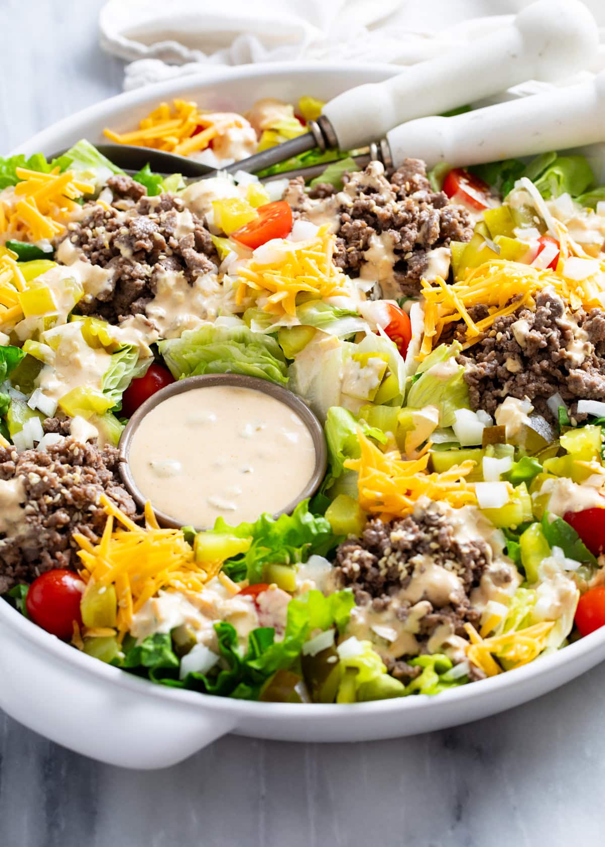 Side view of Big Mac Salad in a white serving bowl with homemade big mac dressing, lettuce, ground beef, onions, pickles, cheese, and tomatoes.